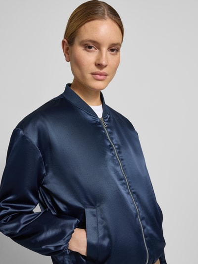 Modelka nosi Regular Fit Blouson mit College-Kragen od Tom Tailor, efekt: jednolity kolor, krój regular fit, kołnierz college, zamek błyskawiczny, długi rękaw, elastyczne zakończenia, wpuszczane kieszenie, wykończenie materiału: satyna, widok z przodu, zbliżenie na górną część ciała.