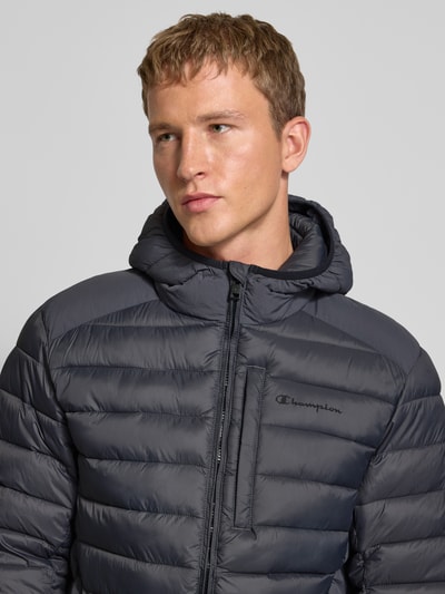 Männermodel draagt Gewatteerd jack met capuchon en logoprint van CHAMPION in donkergrijs, regular fit, ritssluiting, steekzakken, Nahaufnahme Oberkörper. Ruglengte bij maat M: 71 cm, schouderbreedte: 51 cm, borstwijdte: 61 cm, mouwlengte: 69 cm.