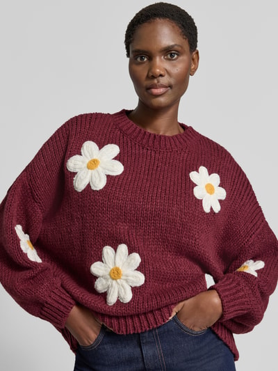 Modelka prezentuje sweter z dzianiny 'Oversized Strickpullover mit floralen Stitchings' od Apricot, bordowy z białymi haftami kwiatowymi, prążkowane wykończenia, okrągły dekolt, dżinsy, widok na górną część ciała z przodu, długość tyłu 62 cm, długość rękawa 43 cm.