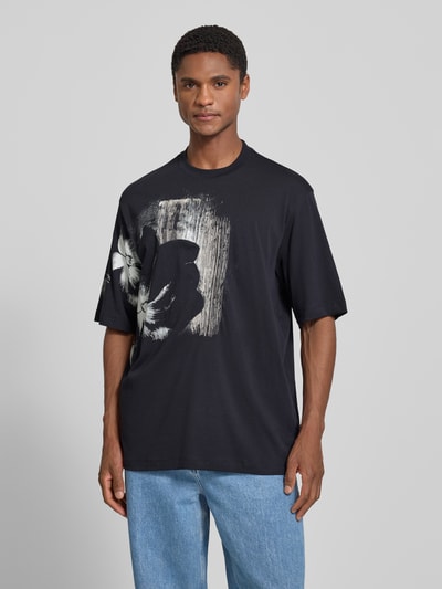 Männermodel trägt Comfort Fit T-Shirt mit Motiv-Print von Armani Exchange, schwarzer Allover-Look, Rundhalsausschnitt, Herren T-Shirt, Ganzkörperansicht von vorne. Rückenlänge bei Größe M: 76 cm.