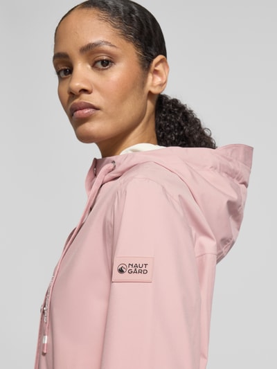 Frauenmodel trägt Jacke mit Ärmelriegeln von NAUTGÅRD in zartem Rosa, glatte Textur, sichtbares Label am Ärmel, Nahaufnahme Oberkörper. Regular Fit, Kapuze mit Tunnelzug, Knopfleiste, Reißverschlusstaschen, Rückenlänge 90 cm, Schulterbreite 45 cm, Brustweite 51 cm, Ärmellänge 62 cm.