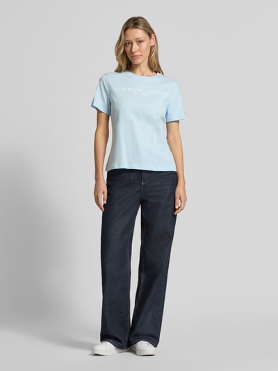Frauenmodel trägt Regular Fit T-Shirt aus reiner Baumwolle von Tommy Hilfiger in Hellblau, unifarben, Logo-Schriftzug, kurze Ärmel, Rundhals, Jersey, Ganzkörperansicht von vorne.