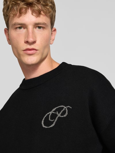 Mężczyznamodel nosi Relaxed Fit Strickpullover aus reiner Baumwolle von PROHIBITED, czarna bawełna, wyhaftowane logo, prążkowane wykończenia, sweter z dzianiny, krój relaxed fit, długość tyłu w rozmiarze M: 64 cm, długość rękawów w rozmiarze M: 59 cm, zbliżenie na górną część ciała.