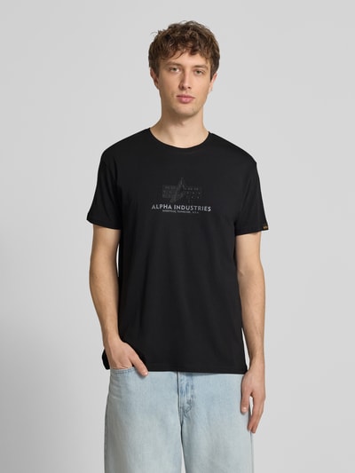 Mężczyzna nosi T-Shirt aus Baumwolle Modell 'BASIC' von Alpha Industries w kolorze czarnym, prosty krój, bawełna, nadruk z logo, okrągły dekolt, widok na cały przód, Ganzkörperansicht von vorne.