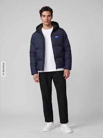Męski model nosi kurtkę pikowaną Regular Fit Steppjacke mit Kapuze Modell 'Bodo2541' von HUGO Blue, dwukolorowy, krój regular fit, kaptur z tunelem, zamek błyskawiczny, naszywka z logo, długi rękaw, elastyczne zakończenie rękawów i dołu, hydrofobowy, boczne wpuszczane kieszenie i kieszeń wewnętrzna, wykonana z popeliny, widok całej sylwetki z przodu, KI-modifiziertes Bild.
