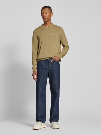 Männermodel trägt Regular Fit Pullover aus Baumwoll-Mix von Lacoste in Beige zu blauen Jeans, Herren Pullover von Lacoste, Ganzkörperansicht von vorne.