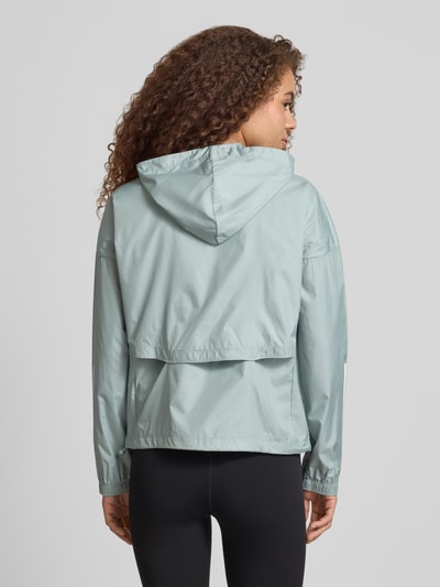 Modelka nosi Windbreaker mit Kapuze und Logo od adidas Sportswear, jasnoszary kolor, lekko błyszcząca tekstura, krój relaxed fit, kaptur, logo, zamek, kieszenie, długość tyłu w rozmiarze S: 58 cm, szerokość ramion w rozmiarze S: 66 cm, szerokość klatki piersiowej w rozmiarze S: 53 cm, długość rękawa w rozmiarze S: 50 cm, widok całej sylwetki od tyłu.
