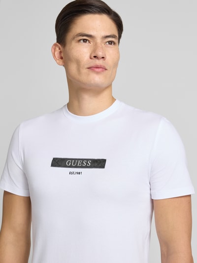 Männermodel trägt T-Shirt met label-detail van Guess in wit, katoen-elastaan mix, regular fit, close-up van de bovenlichaam vooraanzicht.