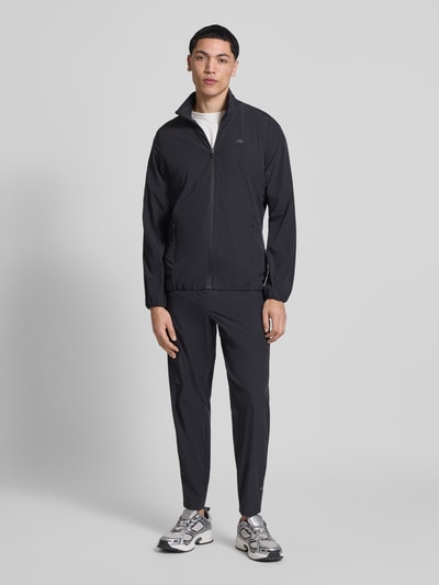 Männermodel trägt Sweatjacke mit Logo-Print Modell 'CANDORA' von Ellesse in Schwarz mit Reißverschluss und Stehkragen, Ganzkörperansicht von vorne. Gerader Schnitt, elastische Abschlüsse, Material-Mix mit Stretch-Anteil, seitliche Reißverschlusstaschen.