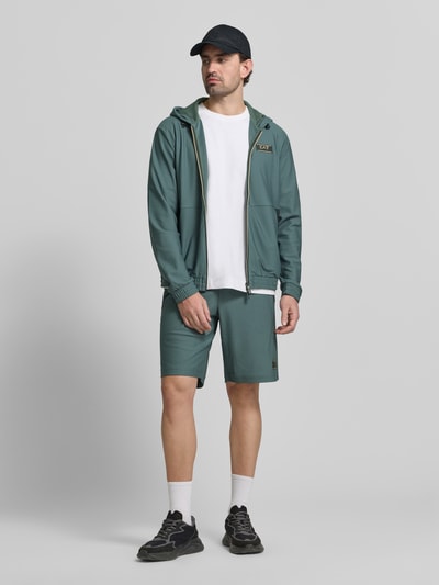 Męski model nosi Regular Fit Shorts mit Strukturmuster od EA7 Emporio Armani, zielony kolor, gumka w pasie, krój regular fit, Szerokość w rozmiarze M: 41 cm, długość nogawki od wewnątrz: 26 cm, widok całej sylwetki z przodu.