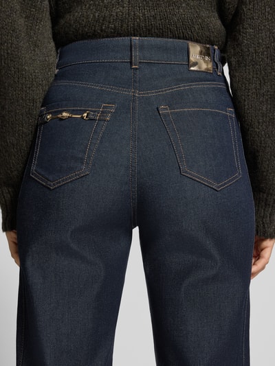 Vrouwenmodel draagt Loose Fit Jeans mit Kettendetail van Marc Cain in donkere denimlook met labelpatch, katoenmix twill, bandplooi, high waist fit, 5-pocketmodel, knoop- en ritssluiting, detailopname rugaanzicht.