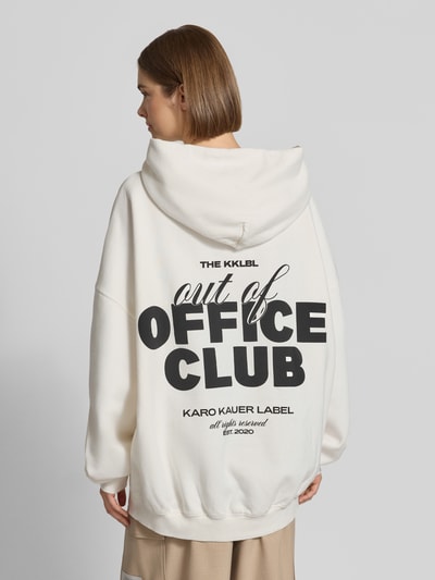 Frauenmodel trägt Oversized Hoodie mit Känguru-Tasche von Karo Kauer, cremefarben, großer Label-Print, lockere Form, Ganzkörperansicht Rückansicht.