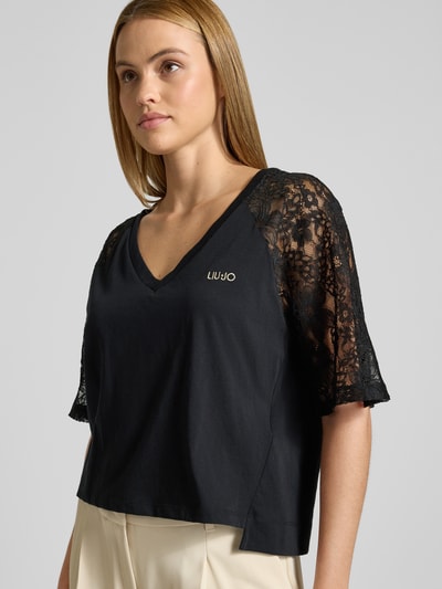 Frauenmodel trägt T-Shirt mit Spitze und Logo-Stitching von LIU JO SPORT in Schwarz aus Reiner Baumwolle, Gerader-Schnitt, V-Ausschnitt, Raglanärmel mit Spitze, Logo-Stitching, Verlängerter Rücken, Rückenlänge bei Größe S: 56 cm, Nahaufnahme Oberkörper.