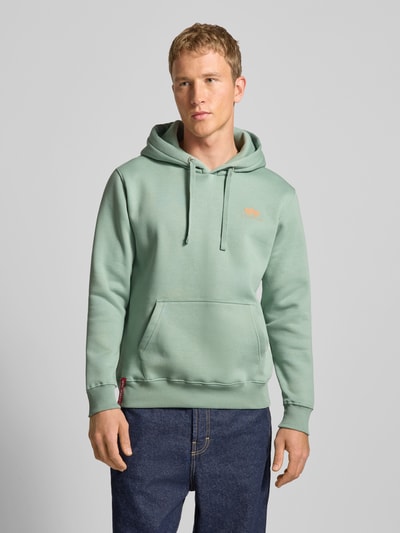 Męski model nosi Hoodie mit Logo und Känguru-Tasche von Alpha Industries, mieszanka bawełny, krój regular fit, długość tyłu 69 cm, długość rękawów 64 cm, jasna zieleń, kieszeń kangurka, widoczny nadruk z logo, gładka faktura, Ganzkörperansicht z przodu.