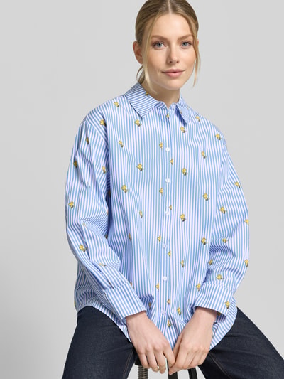 Modelka kobieca nosi Oversized Hemdbluse aus reiner Baumwolle Modell 'JEPPERMINT SNOOPY' von Essentiel Antwerp, niebieskie biało paski z żółtym nadrukiem, motyw Snoopiego, popelina, krój oversize, długie rękawy z mankietami, całościowy wzór, widok na górną część ciała z przodu.