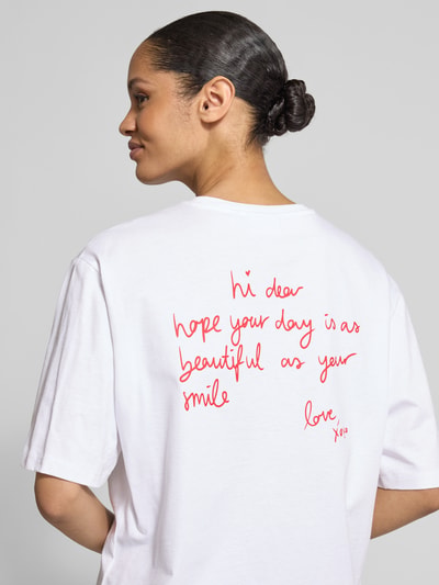Frauenmodel trägt Relaxed Fit T-Shirt mit Motiv-Stitching von comma aus Baumwoll-Elasthan-Mix in Weiß mit rotem Schriftzug, Halbarm-Design, Ganzkörperansicht Rückansicht.