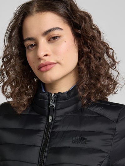 Frauenmodel trägt Steppjacke mit Logo-Stitching Modell 'FUJI' von Superdry in Schwarz, Stehkragen, Reißverschluss, Nahaufnahme Oberkörper. Passform/Schnitt: Gerader-Schnitt, Rückenlänge 62 cm, Schulterbreite 40 cm, Brustweite 50 cm, Ärmellänge 69 cm, Seitliche Reißverschlusstaschen.