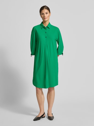 Modelka nosi zieloną sukienkę koszulową z listwą guzikową Knielanges Hemdblusenkleid mit Knopfleiste von Marc Cain, mieszanka materiałów z dodatkiem streczu, prosty krój, długość do kolan, kołnierzyk koszulowy, długość tyłu w rozmiarze 36: 101 cm, szerokość w rozmiarze 36: 54 cm, długość rękawów w rozmiarze 36: 42 cm, widok całej sylwetki z przodu.