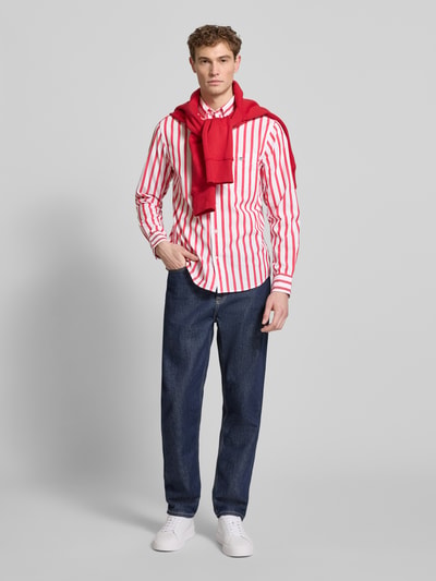 Männermodel trägt Slim Fit Freizeithemd mit Brusttasche von Gant, rot-weiß gestreift, blaue Jeans, rote Baumwollpullover, Ganzkörperansicht von vorne. Herrenhemd aus reiner Baumwolle mit Button-Down-Kragen, Logo-Stitching und Sportmanschetten.