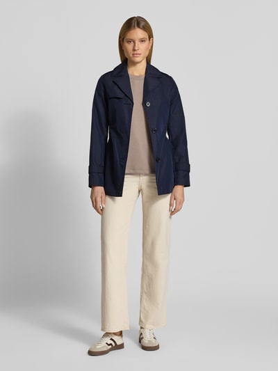 Modelka kobieca nosi Regular Fit Trenchcoat aus Baumwoll-Mix od s.Oliver RED LABEL, jednokolorowy, splot twill, mieszanka bawełny, krój regular fit, kołnierz z połami, listwa guzikowa, pasek w talii, długość krótka, aplikacja z metką, pas na plecach, pagony, długi rękaw z patkami, hydrofobowy, kieszeń wewnętrzna i boczne wpuszczane kieszenie, widok całej sylwetki z przodu.