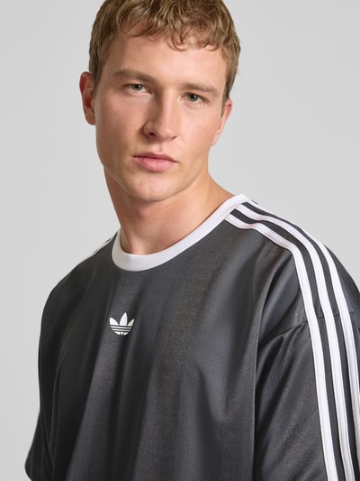 Männermodel trägt Funktionsshirt mit Logo und Rundhalsausschnitt von adidas Originals in Schwarz mit weißen Kontraststreifen, Nahaufnahme Oberkörper. Regular Fit, Rückenlänge bei Größe M 75 cm.