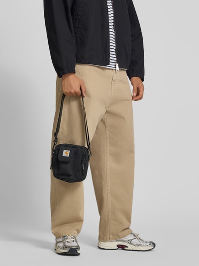 Mężczyzna nosi Crossbody Bag mit Logo-Patch von Carhartt Work In Progress, czarny kolor, regulowany pasek na ramię, główna przegroda z zamkiem błyskawicznym, naszywka z logo, odpinany pasek, szerokość w rozmiarze 1: 17 cm, długość w rozmiarze 1: 18 cm, głębokość w rozmiarze 1: 6 cm, długość paska na ramię w rozmiarze 1: 80 cm, pełny widok z przodu.