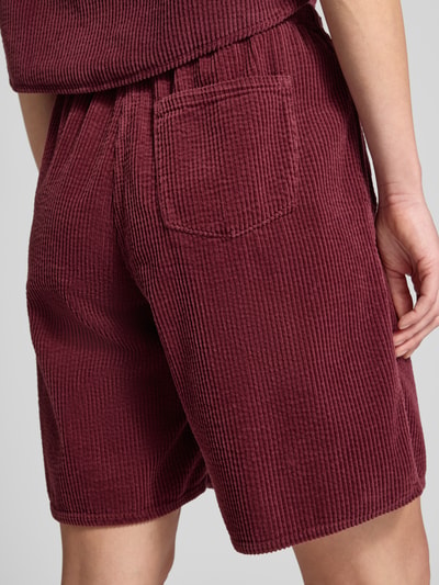 Männermodel trägt Shorts mit elastischem Bund Modell 'PADOW' von American Vintage, bordowy kolor, sztruksowa faktura, luźny krój, kieszeń, czysta wiskoza, Szerokość w rozmiarze S: 37 cm, długość wewnętrzna nogawki S: 20 cm, zdjęcie tyłu w ujęciu całej sylwetki.