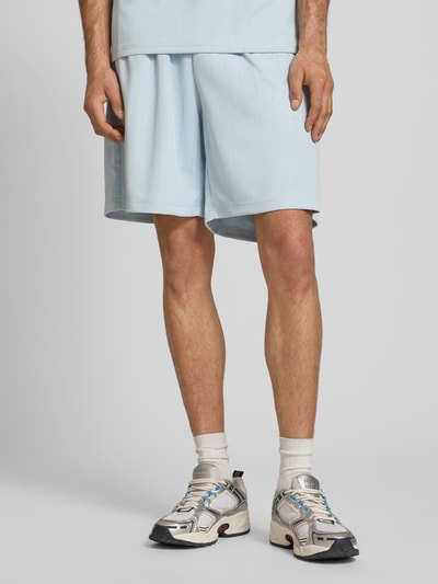Mężczyzna nosi Relaxed Fit Shorts mit elastischem Bund Modell 'Insignia' od KARL KANI, jasnoniebieski, detal z logo, szerokość w rozmiarze M: 37 cm, długość wewnętrzna nogawki: 20 cm, krój relaxed fit, widok z przodu, zdjęcie od kolan w dół.