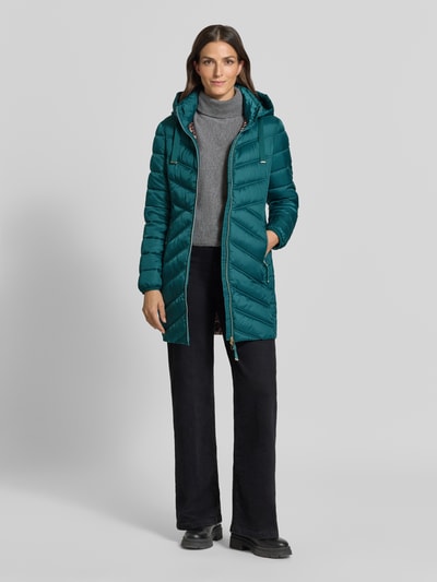 Modelka kobieca ma na sobie Steppmantel mit Zweiwege-Reißverschluss od Christian Berg Woman Selection w kolorze ciemnoturkusowym, krój slim fit, kaptur z troczkiem, kieszenie na zamek, długość tyłu w rozmiarze 36: 83 cm, szerokość ramion w rozmiarze 36: 40 cm, szerokość biustu w rozmiarze 36: 48 cm, długość rękawa w rozmiarze 36: 65 cm, widok całej sylwetki z przodu.