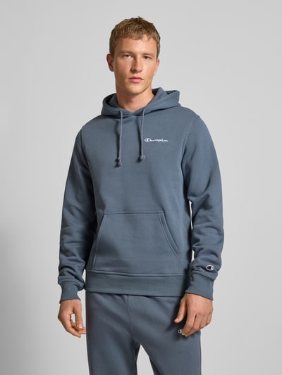 Męski model nosi Hoodie mit Kapuze od Champion, mieszanka bawełny, luźny krój, kaptur, tunel, wyhaftowane logo, kieszeń kangurka, prążkowane wykończenia, długość tyłu 70 cm, długość rękawów 70 cm, kolor niebieskoszary, gładka faktura, całkowity widok z przodu.