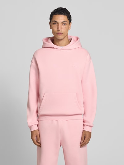 Männermodel trägt Hoodie mit Logo-Print von REVIEW im Baumwoll-Mix, lockere Passform, Kängurutasche, rosa, Ganzkörperansicht von vorne.