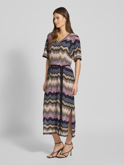Frauenmodel trägt Strickkleid aus Viskose-Mix mit Bindegürtel von Oui, Midikleid mit geradem Schnitt, V-Ausschnitt, 1/2-Arm, buntes Zickzackmuster, Baumwoll-Anteil, Ganzkörperansicht von vorne.