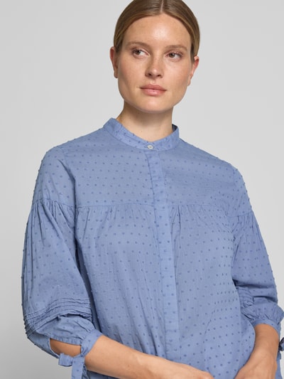 Frauenmodel trägt Regular Fit Bluse mit 3/4-Arm und Knotendetails von Christian Berg Woman aus reiner Baumwolle, Allover-Look, Stehkragen, verdeckte Knopfleiste, dezente Punktstruktur, Nahaufnahme Oberkörper.