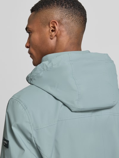 Männermodel trägt Regular Fit Jacke mit Stehkragen von bugatti in Hellblau, glatte Textur, Logo-Detail, verstaubare Kapuze, Detailaufnahme Rückansicht, Zweiwege-Reißverschluss, Eingrifftaschen, elastische Ärmelabschlüsse, Rückenlänge 68 cm, Schulterbreite 42 cm, Brustweite 50 cm, Ärmellänge 65 cm bei Größe 50.
