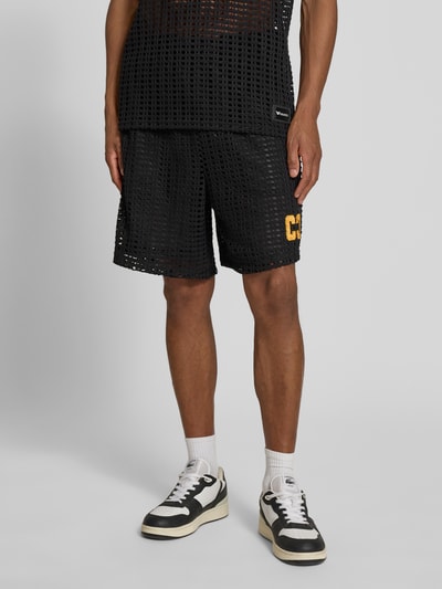 Männermodel trägt Straight Fit Shorts in Loch-Optik von COLUCCI, schwarz mit gelbem Logo-Stitching, elastischer Bund, Ganzkörperansicht von vorne.