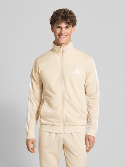 Männermodel trägt Regular Fit Sweat-Anzug mit Kontraststreifen von adidas Sportswear in Beige mit weißen Streifen, unifarben, Ganzkörperansicht von vorne.