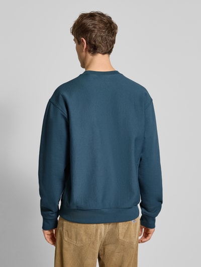 Męski model nosi bluzę Sweatshirt mit Label-Stitching od Carhartt Work In Progress, mieszanka bawełny, luźny krój, prążkowane wykończenia, okrągły dekolt, długość tyłu 69 cm, długość rękawów 65 cm, widok całej sylwetki od tyłu.