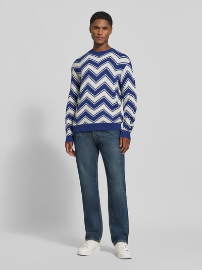 Männermodel trägt Strickpullover aus Schurwolle-Mix mit Rundhalsausschnitt von Armani Exchange, Zickzackmuster in Blau und Weiß, Regular Fit, gerippte Abschlüsse, kombiniert mit Jeans und Sneakers, Ganzkörperansicht von vorne.