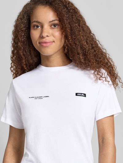 Modelka kobieca nosi T-Shirt mit Logo-Print und Label-Patch od Karo Kauer, biała bawełna, krój straight fit, okrągły dekolt, nadruk logo, metka, długość tyłu w rozmiarze S: 62 cm, zbliżenie na górną część ciała z przodu.