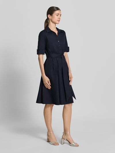 Modelka kobieca nosi Knielanges Hemdblusenkleid mit Bindegürtel od ROBE LÉGÈRE, granatowy bawełniany miks z elastanem, krój slim fit, długość do kolan, kołnierz koszulowy, krótka listwa guzikowa, pasek w talii, rękaw z mankietem, długość tyłu w rozmiarze 36: 104 cm, szerokość w rozmiarze 36: 42 cm, długość rękawa w rozmiarze 36: 30 cm, widok całej sylwetki z przodu.