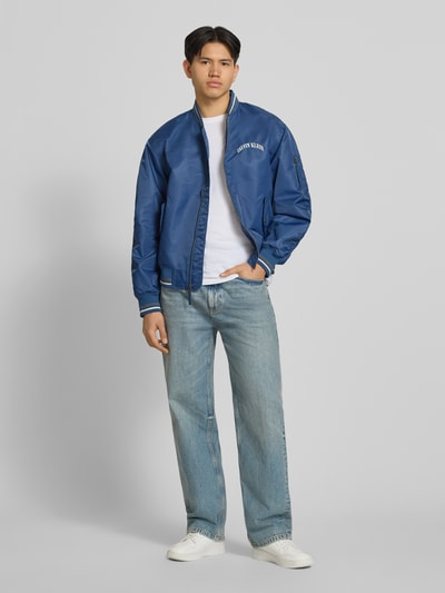 Männermodel trägt Regular Fit College-Jacke mit Label Stitching van Calvin Klein Jeans in look: effen, regular fit, collegekraag, ritssluiting, applicatie met labelstitching, details: contraststrepen, mouwvorm: lange mouw, mouwboord: geribd, rompzoom: geribd, klepzakken. Ganzkörperansicht von vorne.