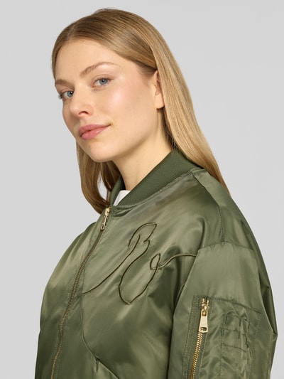 Frauenmodel trägt Bomberjacke mit Zweiwege-Reißverschluss von Original Bombers in Olivgrün, glänzende Textur, Nahaufnahme Oberkörper. Regular Fit, Stehkragen, Stitching, Reißverschlusstaschen, Ärmel mit Zipper, Maße Größe S.