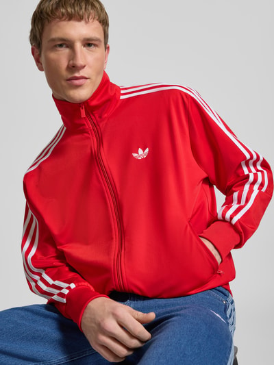 Männermodel trägt rote Sweatjacke mit Logo-Stitching Modell 'FIREBIRD' von adidas Originals, Stehkragen, Reißverschluss, seitliche Reißverschlusstaschen, elastische Abschlüsse, Gerader Schnitt, Nahaufnahme Oberkörper.