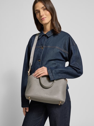 Frauenmodel trägt Handtasche mit Label-Detail von JOOP! in Grau, glatte Textur, goldene Details, abnehmbarer Schulterriemen, Ganzkörperansicht von vorne.