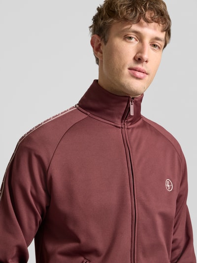 Mannenmodel draagt Trainingsjacke mit aufgesetztem Band auf Raglanärmeln van REVIEW, regular fit, raglanmouwen, bordeauxrood, nahaufnahme bovenlichaam.