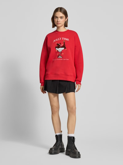 Frauenmodel trägt rotes Sweatshirt mit Motiv-Stitching Modell 'Evie' von LeGer by Lena Gercke, Baumwoll-Mix, Relaxed Fit, Rundhalsausschnitt, schwarze Miniröcke und schwarze Boots, Ganzkörperansicht von vorne.