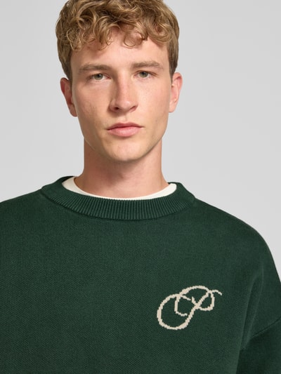 Männermodel trägt Relaxed Fit Strickpullover aus reiner Baumwolle von PROHIBITED in donker groen met labelstitching, Heren, Puur katoen, relaxed fit, losse pasvorm, ribboorden, ruglengte maat M 64 cm, mouwlengte maat M 59 cm, Nahaufnahme Oberkörper.