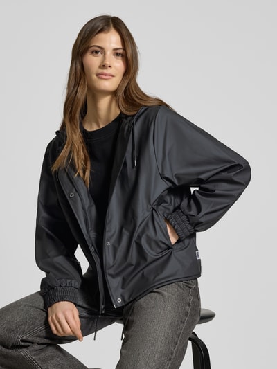 Frauenmodel trägt Jacke mit Abdeckleiste von Rains in Schwarz, gerade Passform, Kapuze mit Tunnelzug, elastische Ärmelabschlüsse, Nahaufnahme Oberkörper. Rückenlänge bei Größe S: 58 cm, Schulterbreite: 43 cm, Brustweite: 55 cm, Ärmellänge: 63 cm.