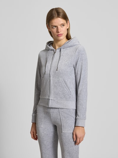 Frauenmodel trägt Sweatjacke mit Label-Stitching Modell 'ROBERTSON' von Juicy Couture aus grauem Material-Mix mit Stretch-Anteil, Regular-Fit, Kapuze mit Tunnelzug, Reißverschluss und Känguru-Tasche, Ganzkörperansicht von vorne.