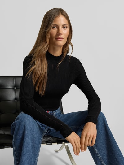 Frauenmodel trägt Slim Fit Pullover mit Woll-Anteil von MYON, schwarz, Stehkragen, Lochmuster, kombiniert mit blauer Jeans, Nahaufnahme Oberkörper.